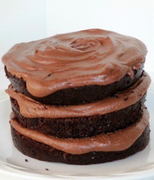 A side view of a three-layer moist chocolate cake and fudge buttercream frosting in between the layers and on top, sitting on a white plate against a white background.