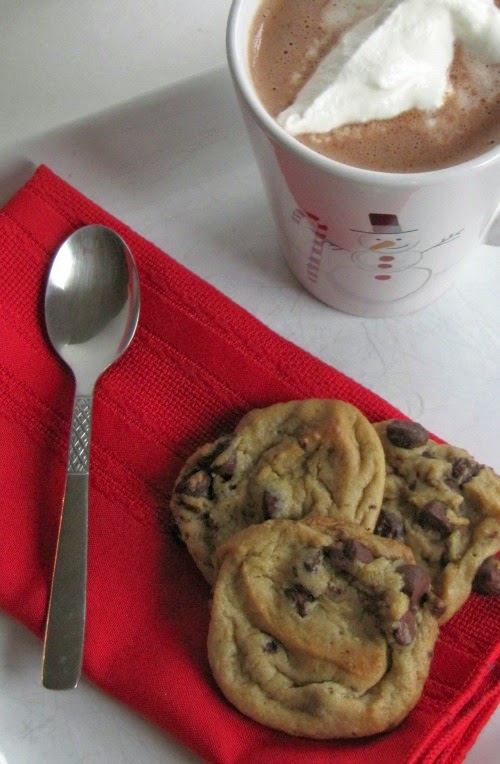 overhead view of three golden brown nestle's toll house cookies on a red napkin with a silver spoon on a white surface next to a white mug of hot cocoa topped with whipped cream