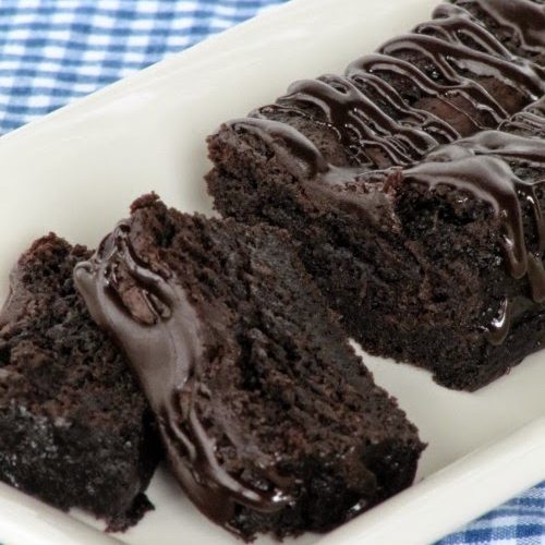 A rectangular white tray set against a blue and white checked cloth holds a partially sliced Nigella Lawson Chocolate Loaf Cake drizzled with melted chocolate.