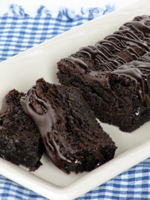 A rectangular white tray set against a blue and white checked cloth holds a partially sliced Nigella Lawson's Chocolate Loaf Cake drizzled with melted chocolate.