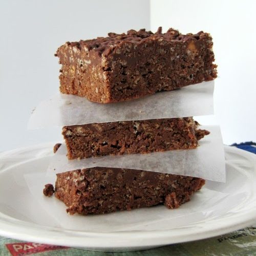 Three dark chocolate nutella rice krispie cereal treats are in a stack separated with pieces of white parchment paper and are sitting on a white plate on a gray marble surface