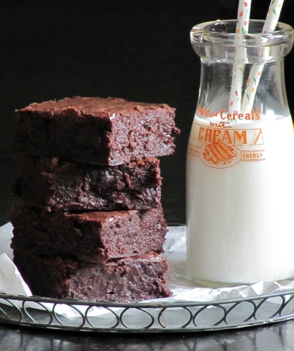 Image showing four dark chocolate Baked Bakery NY aka Oprah's favorite brownies in a stack sitting on white parchment paper next to a glass milk bottle containing milk and two straws.