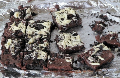 A tin-foil lined pan is full of triple chocolate Oreo bars cut into squares, with Oreo crust, creamy chocolate centers and swirls of dark chocolate and white chocolate plus Oreo chunks on top.