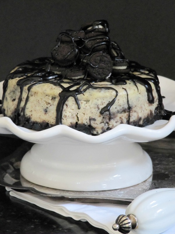 Side view of a large round oreo fudge baked cheesecake made with an Oreo cookie crust, the cheesecake center filled with crushed Oreo cookies, and garnished with a pile of whole oreos and chocolate fudge sauce drizzle.