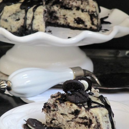 A round oreo fudge baked cheesecake made with an oreo crusst, crushed oreos in the filling, oreos on top and chocolate fudge sauce garnish is on a white cake stand, while a slice of the cheesecake is shown on a white plate in the foreground.