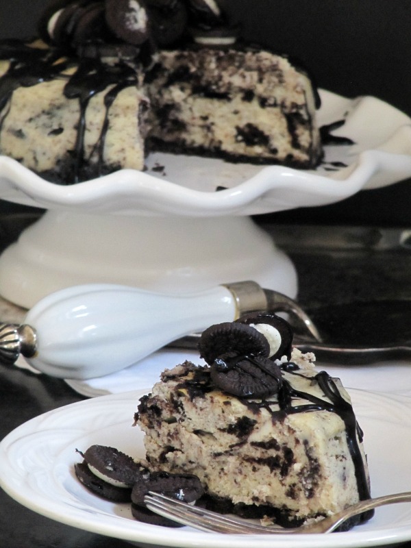 A round oreo fudge baked cheesecake made with an oreo crusst, crushed oreos in the filling, oreos on top and chocolate fudge sauce garnish is on a white cake stand, while a slice of the cheesecake is shown on a white plate in the foreground.