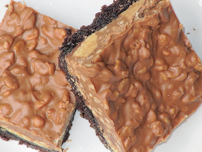 A top view of two peanut butter cup crunch brownies that shows the thick peanut butter cup and rice krispie layer on top of a dark chocolate brownie base on a white surface.
