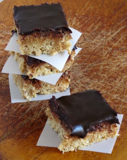 A stack of three browned butter rice krispies peanut butter treats is sitting on a brown surface with a large square on a piece of parchment paper in front of the stack.