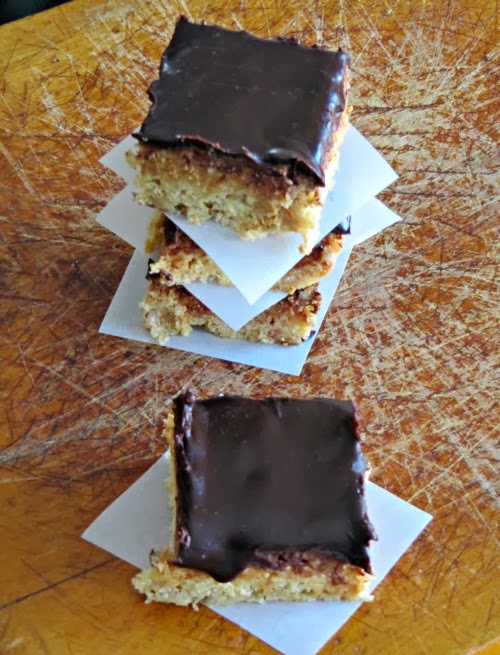 Top view of a stack of three browned butter peanut butter cup rice krispies treats with chocolate ganache topping on a brown surface with a chocolate topped krispie treat sitting in front of the stack.