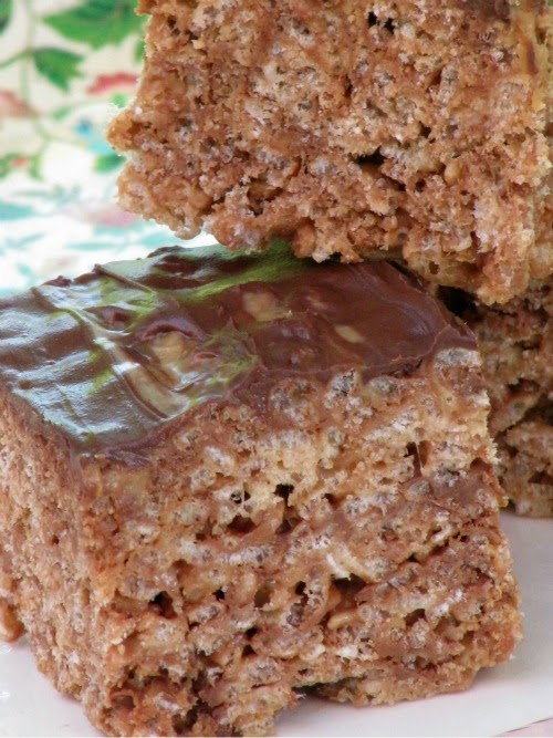 A closeup of a thick Reese's Peanut Butter Cup Rice Krispies treat with a dark chocolate topping with another treat on top of it on a white platter against a colorful floral background.