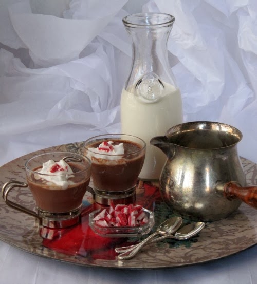 a silver tray against a white background holds two glass cups of Neilsen Massey Peppermint Hot Chocolate topped with whipped cream and crushed mint candies alongside a glass bowl of peppermint candy and two spoons, a silver pitcher of hot chocolate and a tall glass carafe of milk.