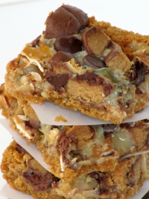 A close up of three pretzel peanut butter cup magic bars in a stack divided with white parchment paper made with graham cracker crumbs, sweetened condensed milk, melted chocolate covered peanut butter cups and pretzels.