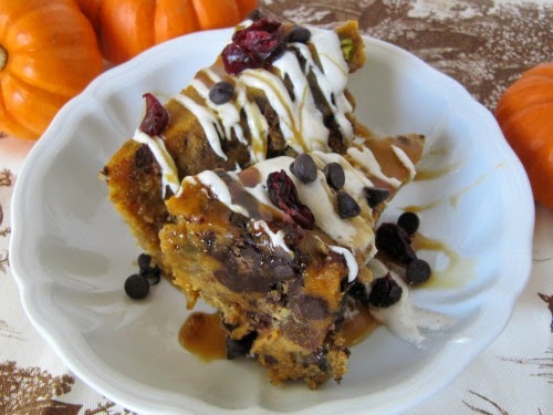 A closeup view of two large pieces of pumpkin bread pudding with Lily's dark chocolate, cranberries and white chocolate drizzle are sitting in a white bowl on a brown and white table cloth along with three orange pumpkins.