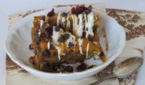 A closeup side view of a white bowl with two pieces of Pumpkin Bread Pudding with Lily's Dark Chocolate, Cranberries and White Chocolate Drizzle itting on a brown and white table cloth along with a serving spoon.