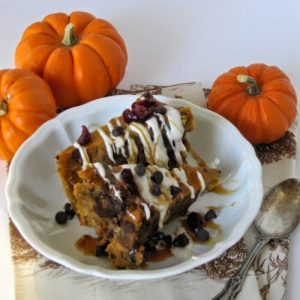 A large white bowl filled with slices of pumpkin bread pudding with dark chocolate chips and cranberries covered with caramel and white chocolate glazes and dark chocolate chips sits on a table along with a spoon and three small orange pumpkins.