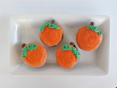 A rectangular white serving tray against a white background is holding four pumpkin patch chocolate cupcakes decorated with orange and green buttercream frosting and tootsie rolls.