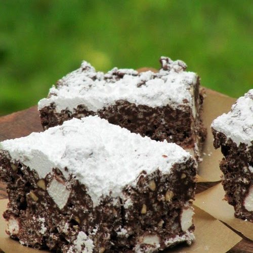 Three puppy chow krispie treats made with chocolate chips, marshmallows, rice krispies and topped with powdered sugar are sitting on brown parchment squares on a wood table against a green background.