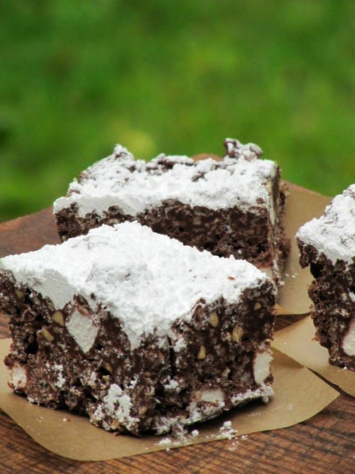 Three puppy chow krispie treats made with chocolate chips, marshmallows, rice krispies and topped with powdered sugar are sitting on brown parchment squares on a wood table against a green background.