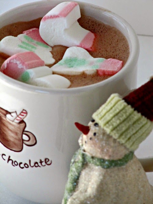 close up overhead view of a white mug filled with creamy raspberry infused hot chocolate topped with green, red and white marshmallwos sitting behind a christmas snowman decoration