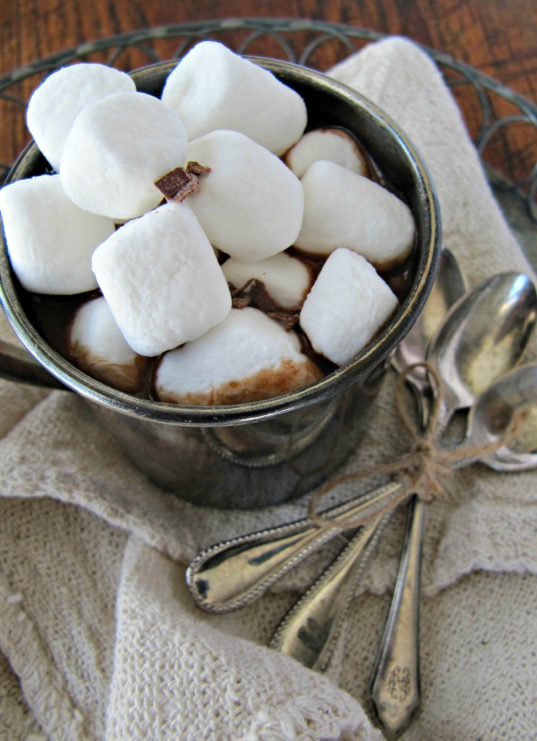 closeup overhead view of a gray mug filled to the brim with marshmallow-topped dark hot chocolate made with real simple magazine's hot cocoa mix recipe. The mug is on a white cloth sitting on a round metal tray along with three silver serving spoon.