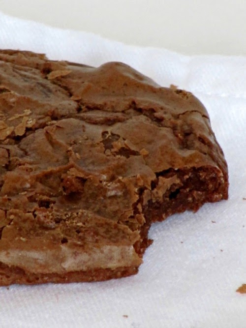 A cloesup of a large chocolate-chip-filled chocolate brownie cookie with a bite taken out of it sitting on a white surface.