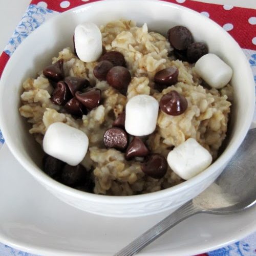 S'mores Oatmeal topped with dark chocolate chips, marshmallows, honey and graham cracker crumbs in a white bowl on a white plate on top of a red, white and blue table covering.