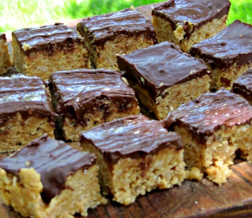 A group of Toblerone Cereal Treats made with peanut butter and rice krispie cereal and topped with melted Toblerone mini chocolate almond candies are sitting on a wood surface against a green background.