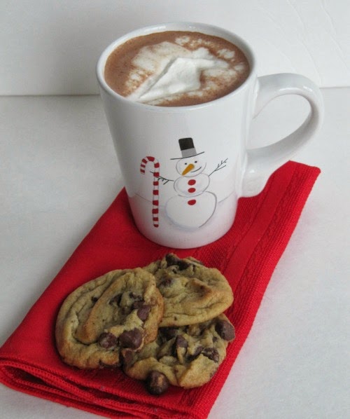 front view of a white surface with a red cloth napkin holding three light brown Nestle's toll house cookies along with a white snowman mug filled with hot chocolate topped with whipped cream.