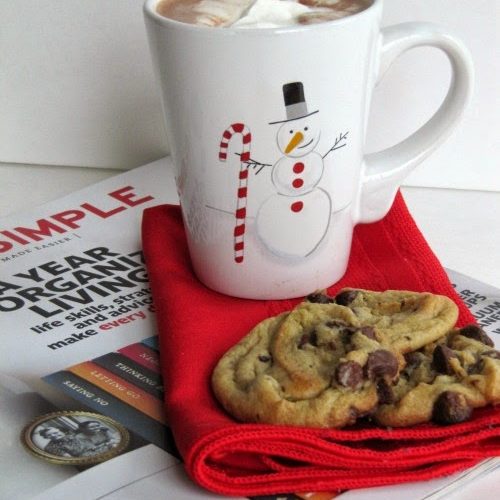 Side view of a white surface with a simple Magazine on which is a red napkin holding three nestle's toll house cookies and a white christmas mug filled with hot cocoa and whipped cream topping