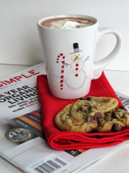 Side view of a white surface with a simple Magazine on which is a red napkin holding three nestle's toll house cookies and a white christmas mug filled with hot cocoa and whipped cream topping.