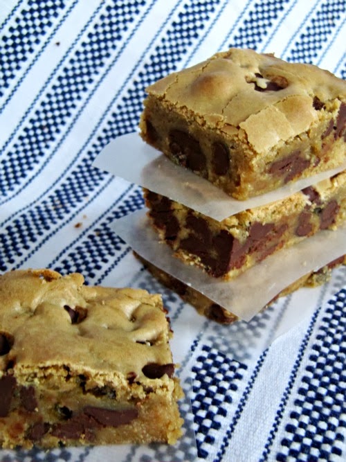 An angled side view of a stack of three triple chocolate blondies brownies and a single blondie sitting to the side, all of which have crunchy tan tops and centers swirled with melted milk, semi-sweet, and dark chocolae chips. The bars are sitting on a blue and white striped woven table cloth.