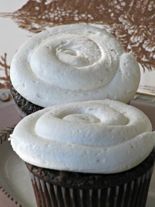 A closeup photo of two chocolate cupcakes frosted with a thick swirl of white marshmallow buttercream frosting for chocolate cupcakes, shown against a vintage floral brown background.