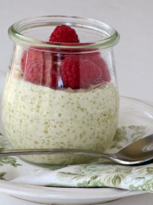 A glass jar of creamy white tapioca pudding with fresh raspberies on top is sitting on a tan and white floral napkin on a white plate with a silver spoon.