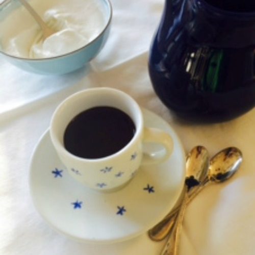 A white tablecloth holds a blue and white cup and saucer filled with dark Viennese hot chocolate next to a bowl of whipped cream, two silver spoons, and a black pitcher of hot chocolate.
